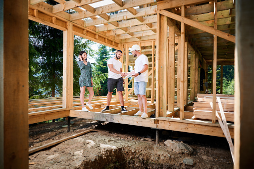 Man with detailed plan in hand showcases to couple the process of constructing wooden framed house near the forest. Men firmly grasp each other's hands in a gesture of mutual respect.