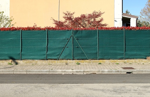 House fence made of net and green tarpaulin. Plants and wall on behind, concrete tile sidewalk and street in front. Background for copy space.