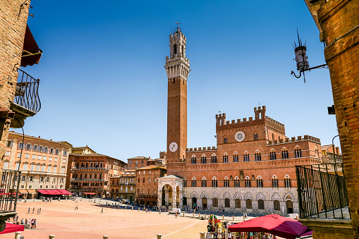 A suggestive glimpse of Piazza del Campo, in the medieval heart of Siena, in Tuscany, with the majestic Torre del Mangia (Mangia tower) and the Palazzo Pubblico. Built in 1290 as the seat of the Council of Nine and of the executive power of the city, the majestic Palazzo Pubblico is still the seat of the municipal government. The Torre del Mangia was built between 1338 and 1348 on a project by Giovanni Balduccio, known as Mangia, becoming the symbol of the political and military power of Siena compared to its rival Florence. Center of the city life since 1169, Piazza del Campo or simply Campo is one of the most beautiful and famous squares in the world for its particular shell shape. In this space every year the seventeen historical neighborhoods of Siena compete in the Palio, one of the oldest horse races in the world. Siena is one of the most beautiful Italian cities of art, in the heart of the Tuscan hills, visited for its immense artistic and historical heritage and for its famous popular traditions. Since 1995 the historic center of Siena has been declared a World Heritage Site by UNESCO. Wide angle image in High Definition quality.