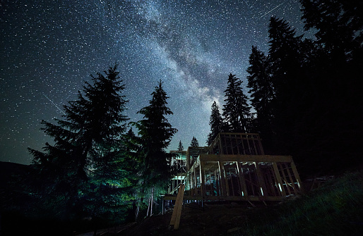 Nighttime photograph of residential wooden frame house under construction near the forest against a starry sky and Milky way. Start of construction of cozy home or cottage on the edge of the forest.