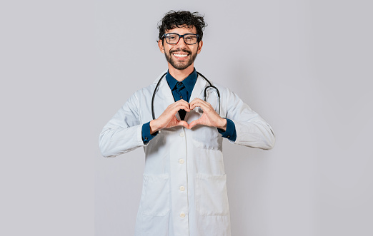 Young doctor making heart shape isolated. Handsome doctor making heart gesture with hands. Happy doctor pointing fingers down