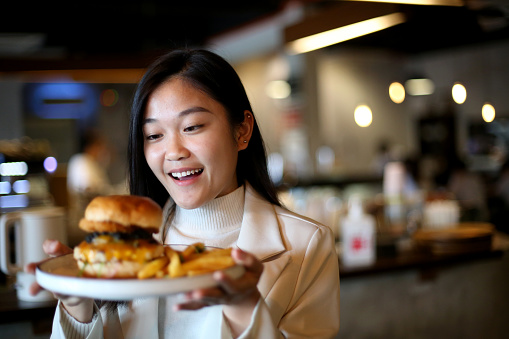 An Asian young woman is holding a hipster burger with fries in the café