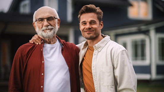 Portrait of a Happy Senior Father or Grandparent Posing Together with His Handsome Adult Son. Family Members Embrace Each Other, Standing, Looking at Camera and Smiling.