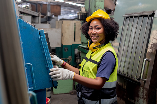 engineering female african american happy smiling workers wear soundproof headphones and yellow helmet holding tablet working at operating CNC machine. work factory industrial concept