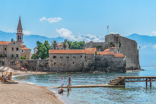Budva, Montenegro - June 17, 2021: Plaza Ricardova Glava Beach near the walls of the Old Town of Budva. People rest on the sandy coast of the Adriatic Sea with a fortress among the blue water