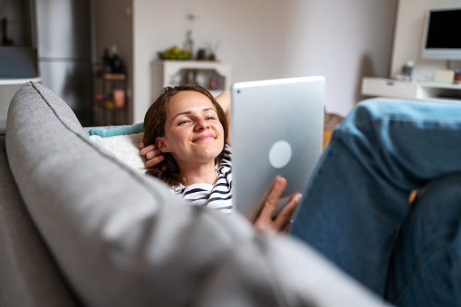 Woman watching vlog on internet and smiling while lying on coach at home