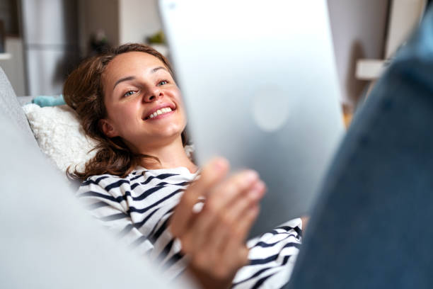 istock Woman smiling and using her digital tablet at home lying down on sofa. 1481354352