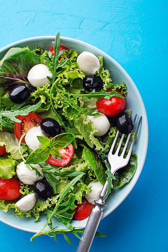 Caprese Italian or Mediterranean salad. Green Salad with mozzarella, black olives and tomato on blue table background close up