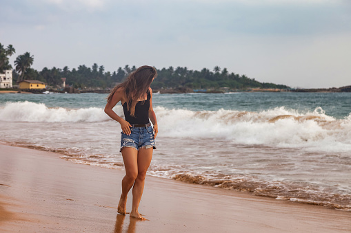 Young woman in denim shorts walking along sandy beach at ocean waves background, summer vacation. Traveler lady walks on tropical seacoast, relaxing lifestyle. Tourist holiday concept. Copy text space