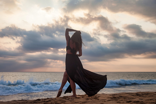 Rear view elegant woman posing on sandy beach at sunset ocean background; summer vacation. Back view full length lady stands on tropical seacoast; sunset. Tourist holiday concept. Copy ad text space