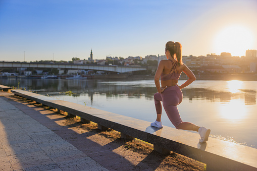 Woman in sports outfit stretching outdoors before sports activities, standing in lunge position on platform by the city river at sunset