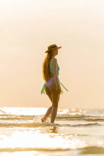 Photo of Beautiful caucasian woman in bikini and hat wandering at the beach during an amazing sunset moment.