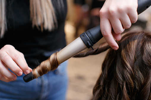 Using hot curler and making curls on brunette customer's hair strands, changing hairstyle