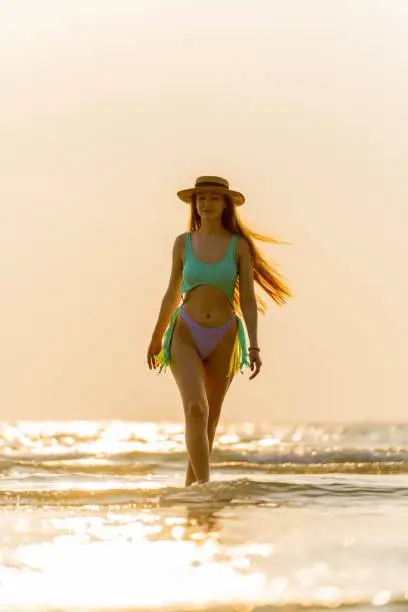 Photo of Beautiful caucasian woman in bikini and hat wandering at the beach during an amazing sunset moment.