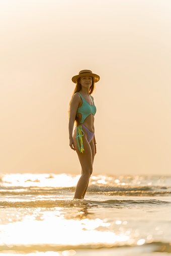 Portrait photos of a beautiful caucasian woman in bikini and hat wandering at the beach during an amazing sunset moment.