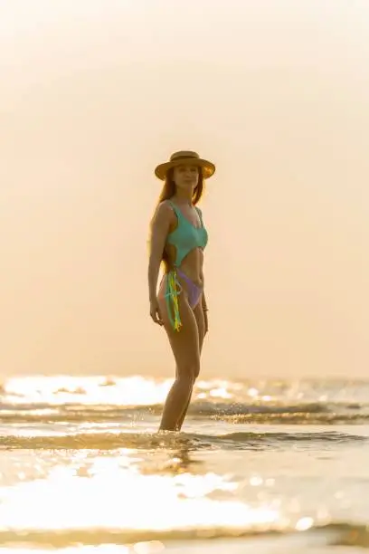 Photo of Beautiful caucasian woman in bikini and hat wandering at the beach during an amazing sunset moment.