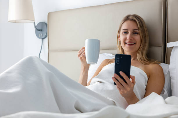 istock smiling young woman laying in bed and using phone while drinking morning coffee at home bedroom 1481364880