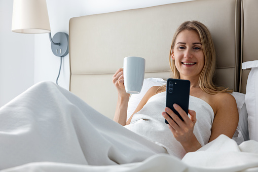 smiling young woman laying in bed and using phone while drinking morning coffee at home bedroom