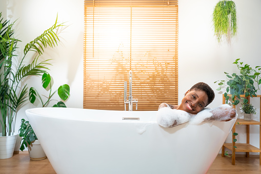 Portrait of an attractive African woman relaxing in bathtub at cozy home bathroom looking at camera smiling happy. Real people wellness concept. High quality photo