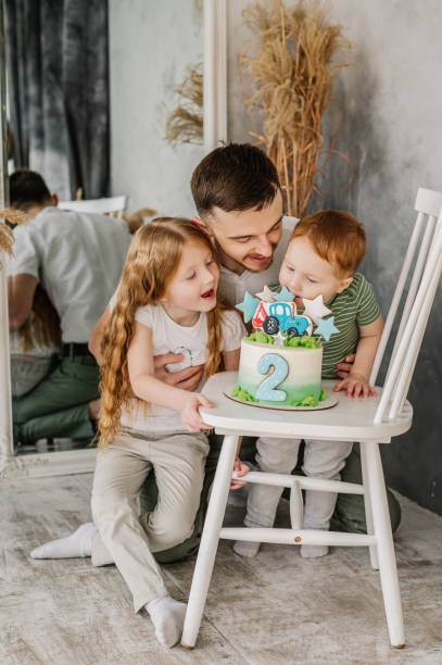 feliz familia soplando velas en el pastel y divirtiéndose celebrando la fiesta de cumpleaños de 2 años. padre abrazando a los niños. recepción de cumpleaños. papá e hija felicitan a su hijo por su cumpleaños en el estudio. come pastel. - 2 3 years children only group of people enjoyment fotografías e imágenes de stock