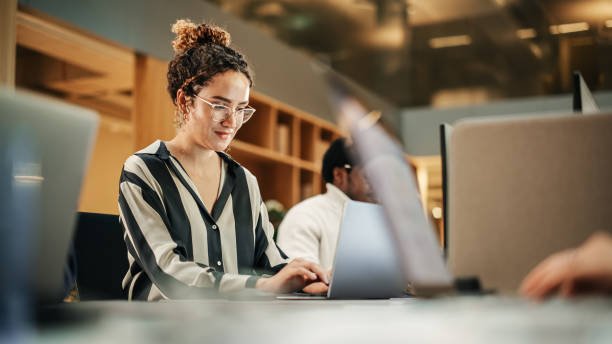 istock Diverse Group of Creative Colleagues Working on Laptops in Modern Office. Hispanic Female Graphics Designer Smiling and Typing. Colleagues Working on Projects in Background. 1481368984