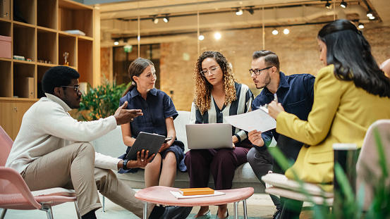 Group of People from Multiple Ethnicities Working on Problem Solving Using Notes, Laptop and Tablet in a Meeting Room at the Office. Teammates Giving Constructive Feedback on Eachother's Projects