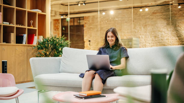Portrait of Caucasian Creative Young Woman Working on a Laptop in Bright Casual Office. Female Client Coordinator Smiling When Receiving Positive Feedback From Clients. Employees Walking in Background retrato de una joven creativa caucásica trabajando en una computadora portátil en una oficina informal y brillante. coordinadora de clientes sonriendo cuando recibe comentarios positivos de los clientes. empleados caminando en segundo plano