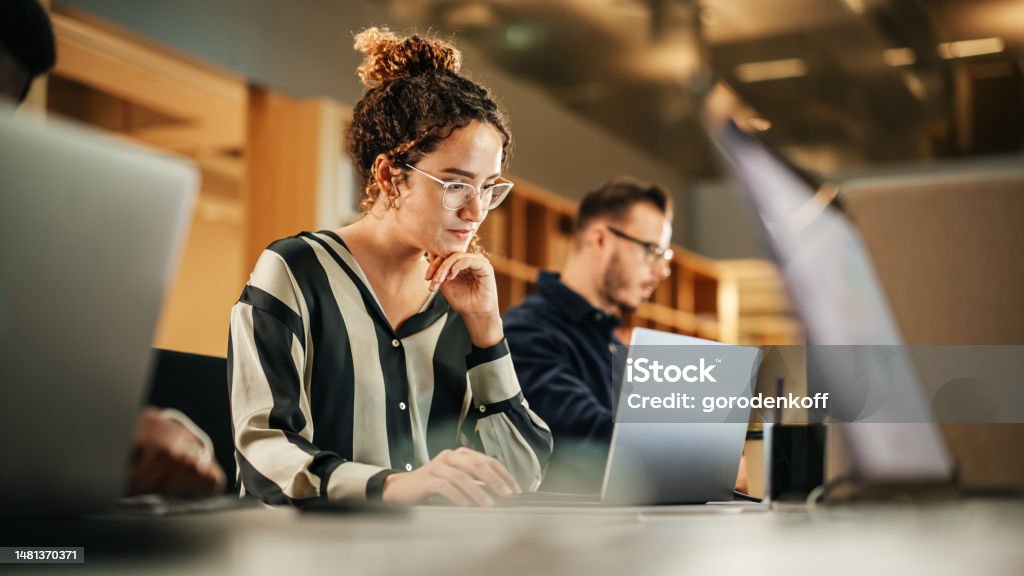 Portrait of Enthusiastic Hispanic Young Woman Working on Computer in a Modern Bright Office. Confident Human Resources Agent Smiling Happily While Collaborating Online with Colleagues. Office Stock Photo