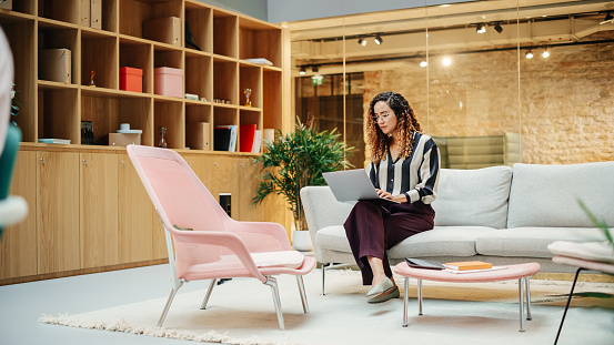 Portrait of Hispanic Creative Young Woman Working on a Laptop in Casual Office. Female Team Lead Smiling While Checking her Team Performance Data. Focused Hard Worker. Wide Shot