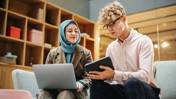 istock Portrait of Two Creative Colleagues Smiling and Discussing Work Using Laptop and Tablet in Office. Young White Male Marketing Intern Consulting Female Caucasian Creative Director. 1481370580