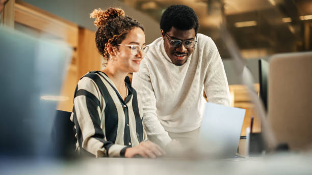 porträt von zwei kreativen kollegen, die einen laptop benutzen, um arbeitsprojekte im büro zu besprechen. young black technical support specialist hilft weiblicher kundenbeziehungskoordinatorin. teamwork-konzept - half staff stock-fotos und bilder