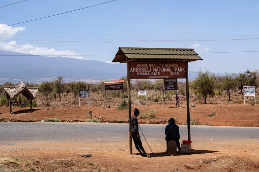 Kenya, Africa - March 5, 2023: Various signs directing drivers to lodges and camps near Amboseli National Park. Two villagers sit by the sign