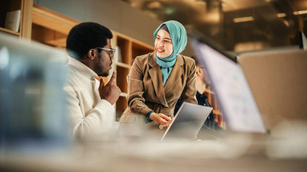 zwei mitarbeiter, die zusammenarbeiten und ihre leistung in einem modernen büro feiern. muslimische trainerin, die einen laptop benutzt, um einen schwarzen männlichen kundendienstmitarbeiter an bord zu bringen und seine frage zu beantworten. - religiöse kleidung stock-fotos und bilder