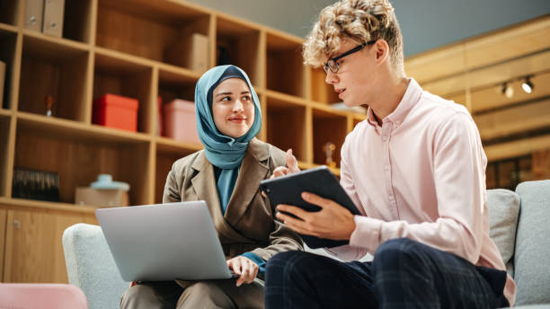 istock Two Creative Colleagues Discussing and Pointing at a Laptop in Modern Casual Office. Caucasian Male Sales Manager Discussing Project Plan on Tablet with Muslim Female Legal Assistant. 1481370821