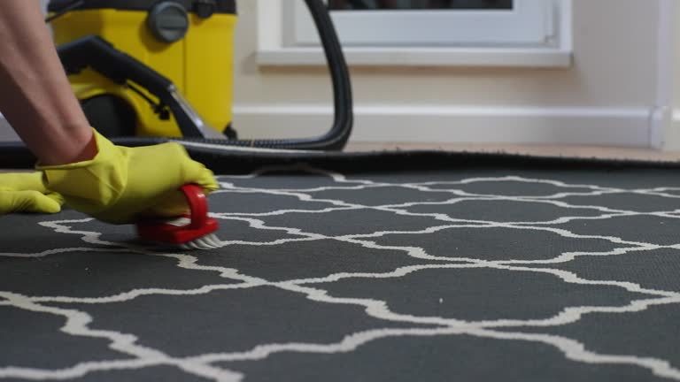 Side view cropped shot of unrecognizable janitor male in yellow rubber gloves using detergent and brush cleaning carpet at home on background of vacuum cleaner.