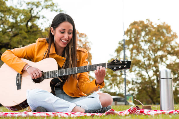 latin young woman sitting in the park smiling and playing guitar stock photo