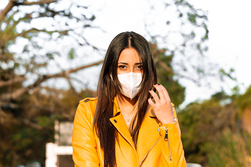 portrait young latin woman, with yellow jacket, in the park at sunset with a mask looking at the camera
