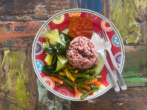 Healthy vegan nasi campur served with red rice, stir fried vegetables, tempeh and hot chili sauce on rustic colorful table.  Canggu, Bali, Indonesia.
