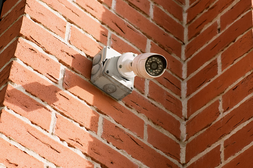 Small white CCTV camera on the corner of the facade of a multi-storey brick building. Security surveillance camera with a broken plastic case.