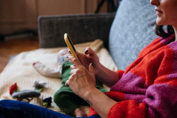 Photo of An Unrecognizable Beautiful Businesswoman Texting On Her Mobile Phone While Sitting On The Sofa Next To Her Baby