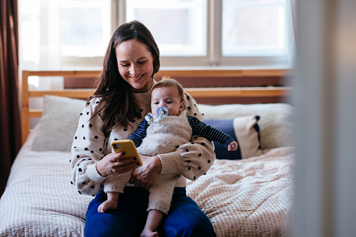 A smiling Caucasian entrepreneur using her smartphone while taking care of her newborn child. She is sitting on the bed in her bedroom.