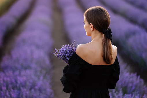 Back view of young romantic woman with healthy natural beauty skin holding bouquet in lavender field looking at camera and smiling. Beautiful girl wearing black dress and bow on hair enjoying view