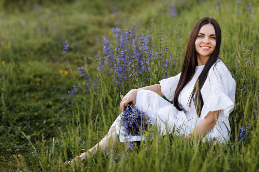 Young smiling beautiful brunette woman in white dress is sitting in green meadow with lupine flowers in spring. Happy girl looks at camera. Women's day. Summertime. Romantic mood. Nature concept.