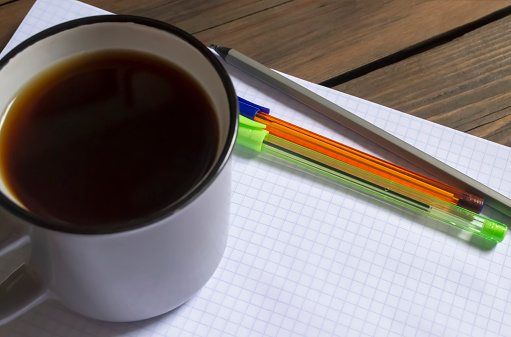 A cup of coffee next to the notepad and pens. On the table is a plaid notebook, three pens and a cup of black coffee. Americano during his studies.