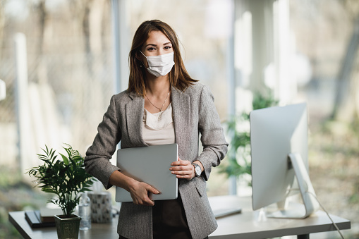A successful young business woman with surgical mask holding laptop and standing in her office. Looking at camera.