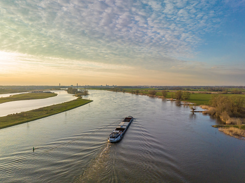 Panoramic aerial view on a freight ship sailiing on the river IJssel during a springtime sunset in Overijssel. The flow of the river is leading towards the setting sun in the distance while lights are popping up in the city at the end of a beautiful springtime day.