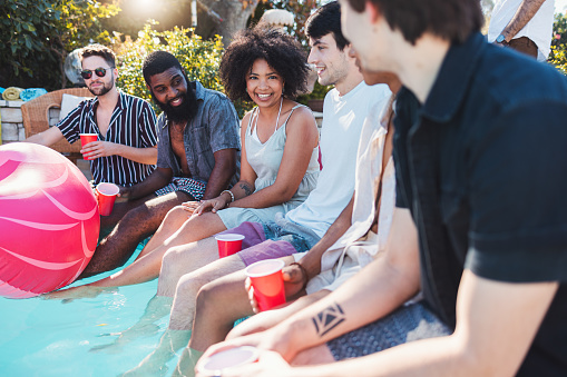 Holiday, drinks and people speaking in a pool at a summer party, celebration or event at a home. Diversity, vacation and friends talking, having fun and bonding by the swimming pool while drinking.