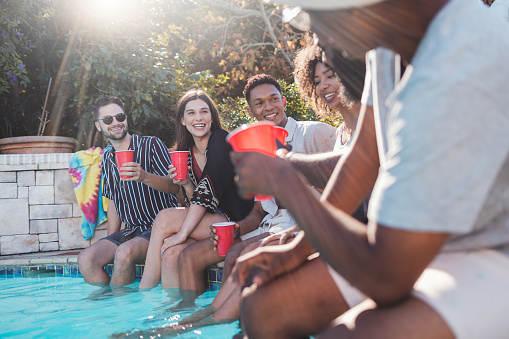Vacation, drinks and friends speaking in a pool at a summer party, celebration or event at a home. Diversity, happy and people talking, having fun and bonding by the swimming pool while drinking.