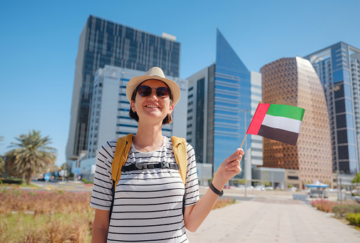 Happy young asian female traveler with backpack and hat with UAE flag against scenic skyscrapers in Abu Dhabi. Immigration, student education and arab emirates citizenship concept