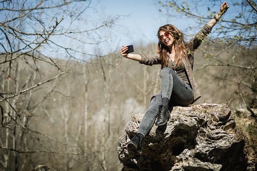 A cheerful young woman sitting on the top of the mountain cliff, making a selfie and enjoying the outdoors.